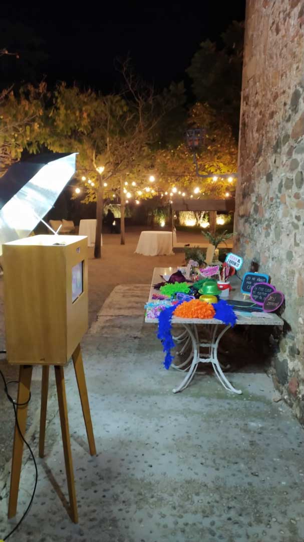 Fotomatón de lado orientado hacia una pared de piedra usada como photocall, con una mesa de atrezo al fondo, incluyendo gorros, gafas y pelucas. En el fondo, mesas y luces decorativas indican la celebración de una boda en Valencia con guirnaldas de colores.
